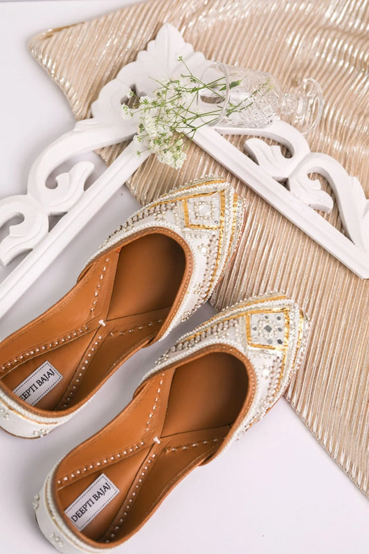Pair of white and gold embellished handcrafted jutti shoes on decorative frame with glass vase and white flowers