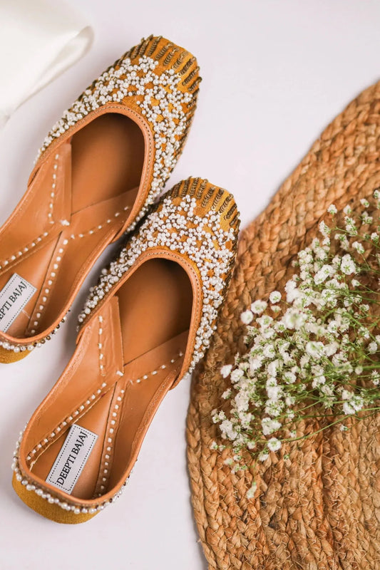 Handcrafted women's jutti in mustard with intricate white pearl beadwork on leather soles, placed beside dried white flowers on woven mat