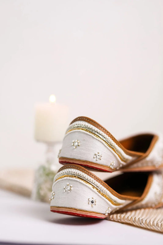 Close-up of elegant white women's jutti with pearl and gold bead embroidery, lit candle in background