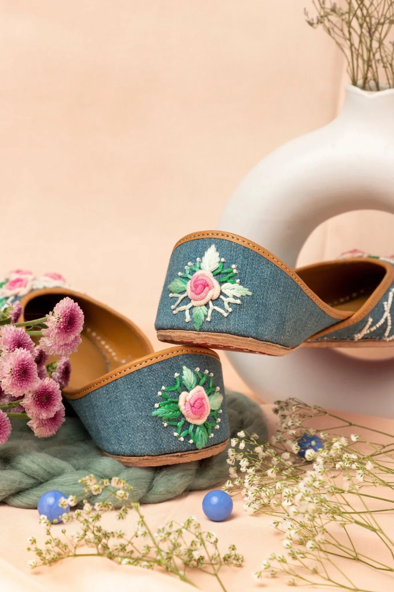 Handcrafted blue denim jutti for women with pink floral embroidery, styled with flowers and white ceramic decor