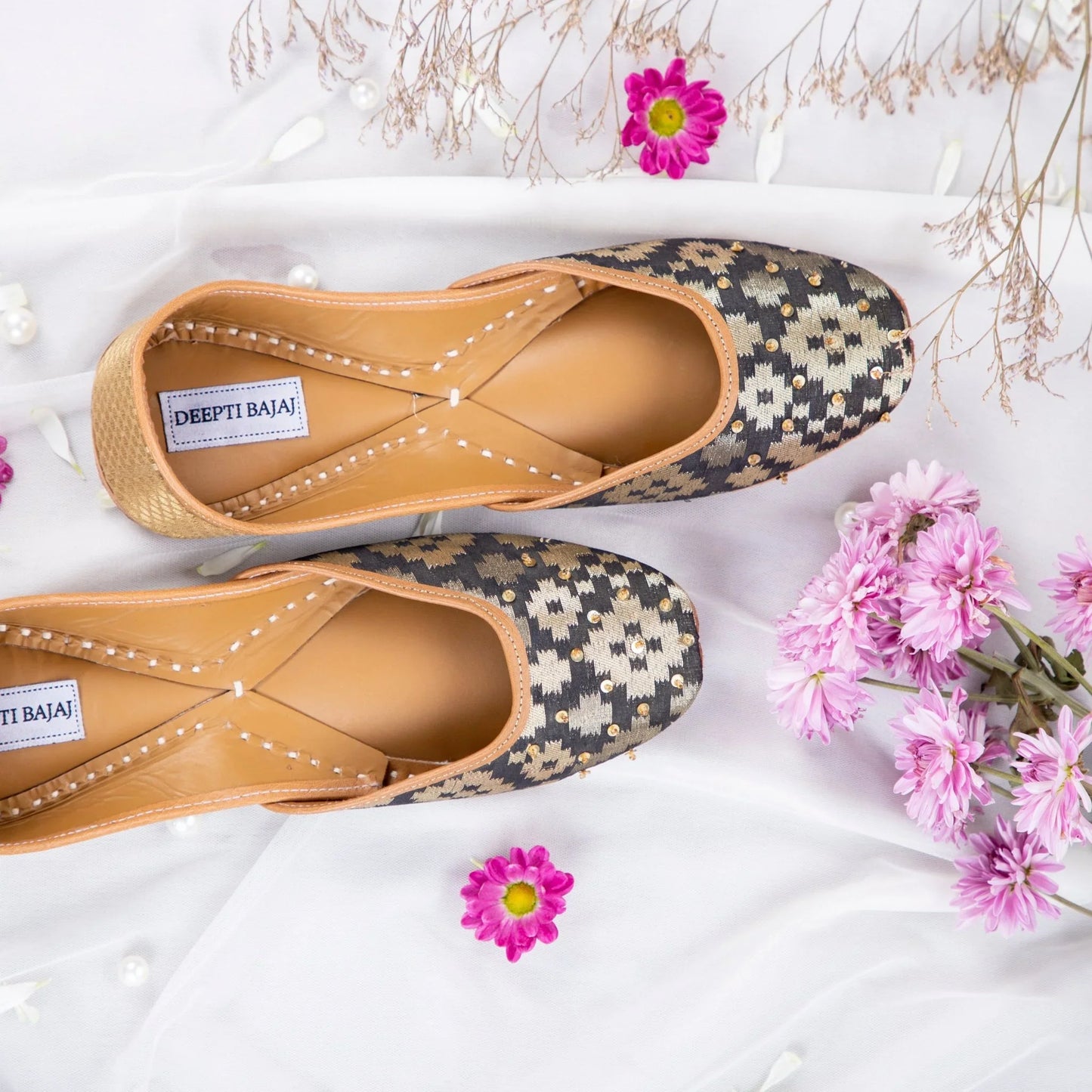 Handcrafted women's jutti by Deepti Bajaj with black and gold ethnic pattern, adorned with small beads, displayed on white fabric with pink flowers