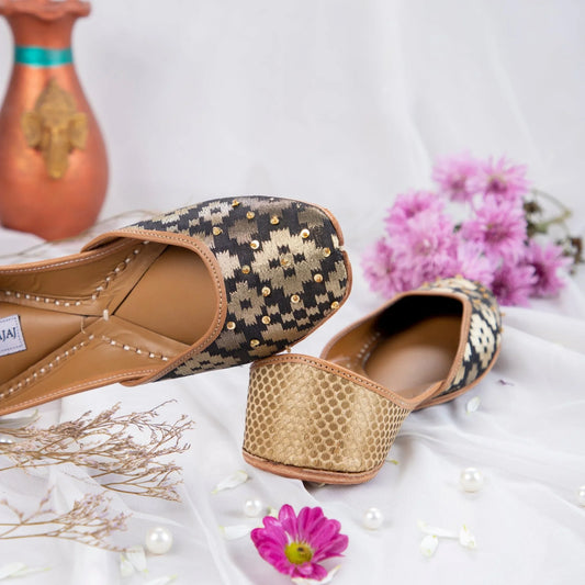 Handcrafted women's jutti with black and gold brocade, gold embellishments, displayed on white fabric with purple flowers and pearls