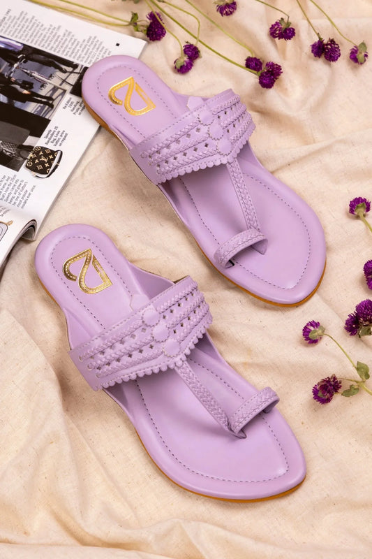 Pair of lavender handcrafted women's jutti sandals with intricate braided design on beige fabric, purple flowers, and magazine