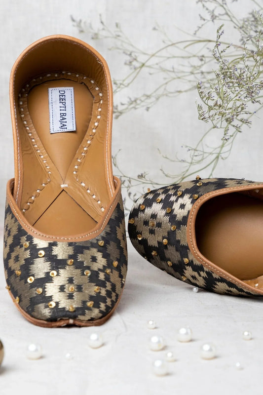 Handcrafted women's jutti with black and gold geometric pattern, embellished with sequins on beige background
