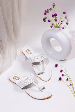 Pair of white handcrafted jutti sandals with perforated design beside a modern round white vase with purple flowers
